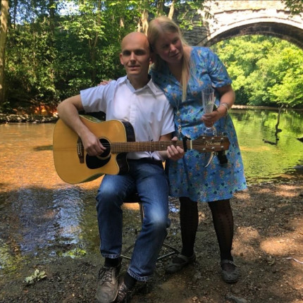 Photo of Vicky (right) standing next to her husband (left) who is sat on a chair