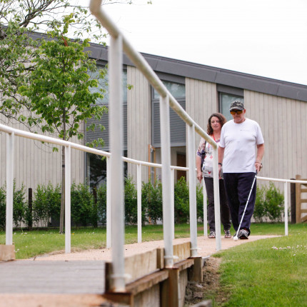 Veteran walks along walkway using long cane, accompanied by rehabilitation officer