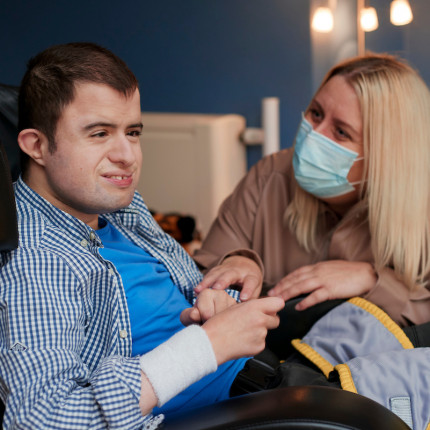 A young man using a wheelchair smiles at the camera while his care worker stands beside him