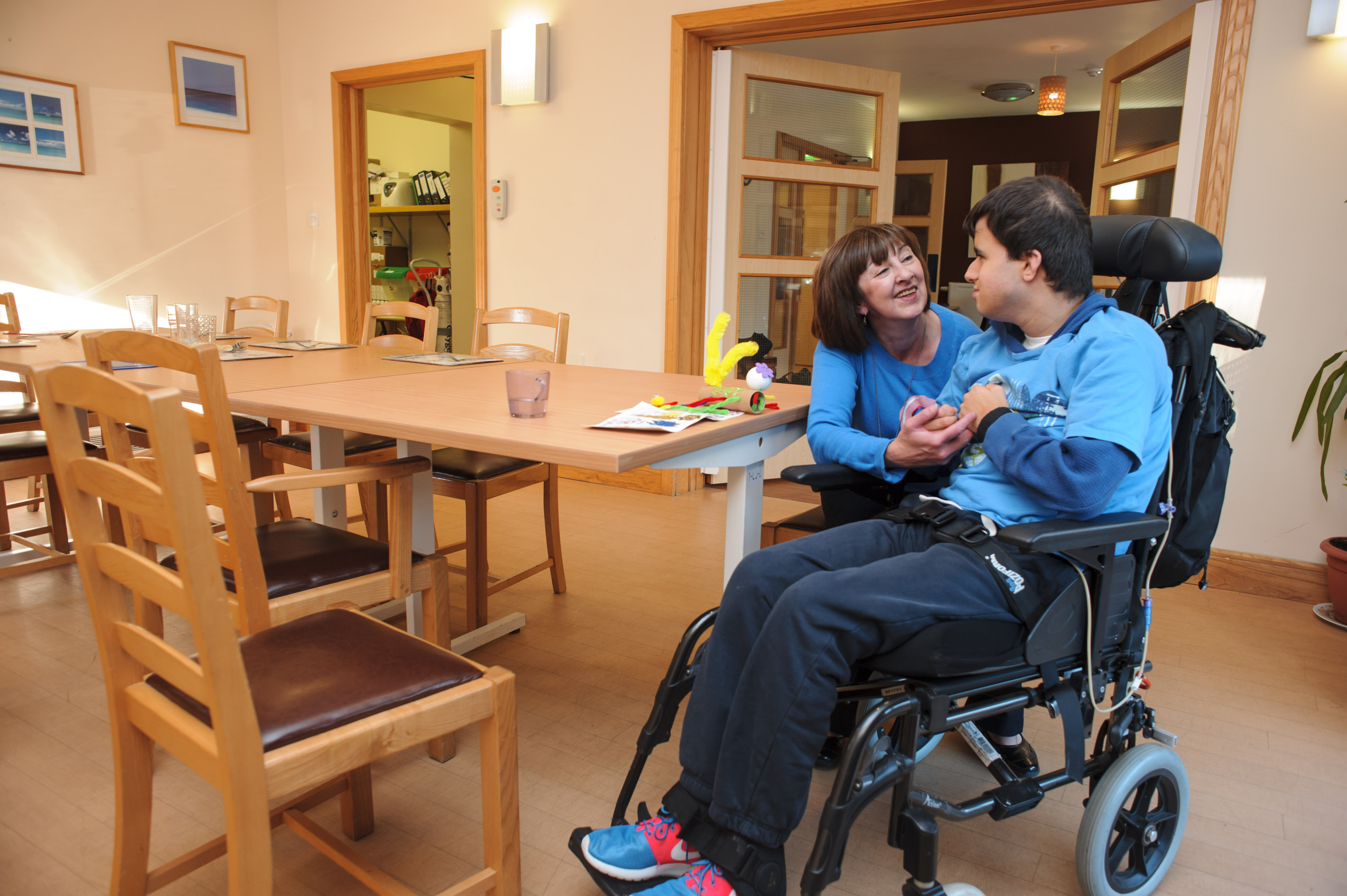 Pupil seated in wheelchair at dining room table smiles at care staff member