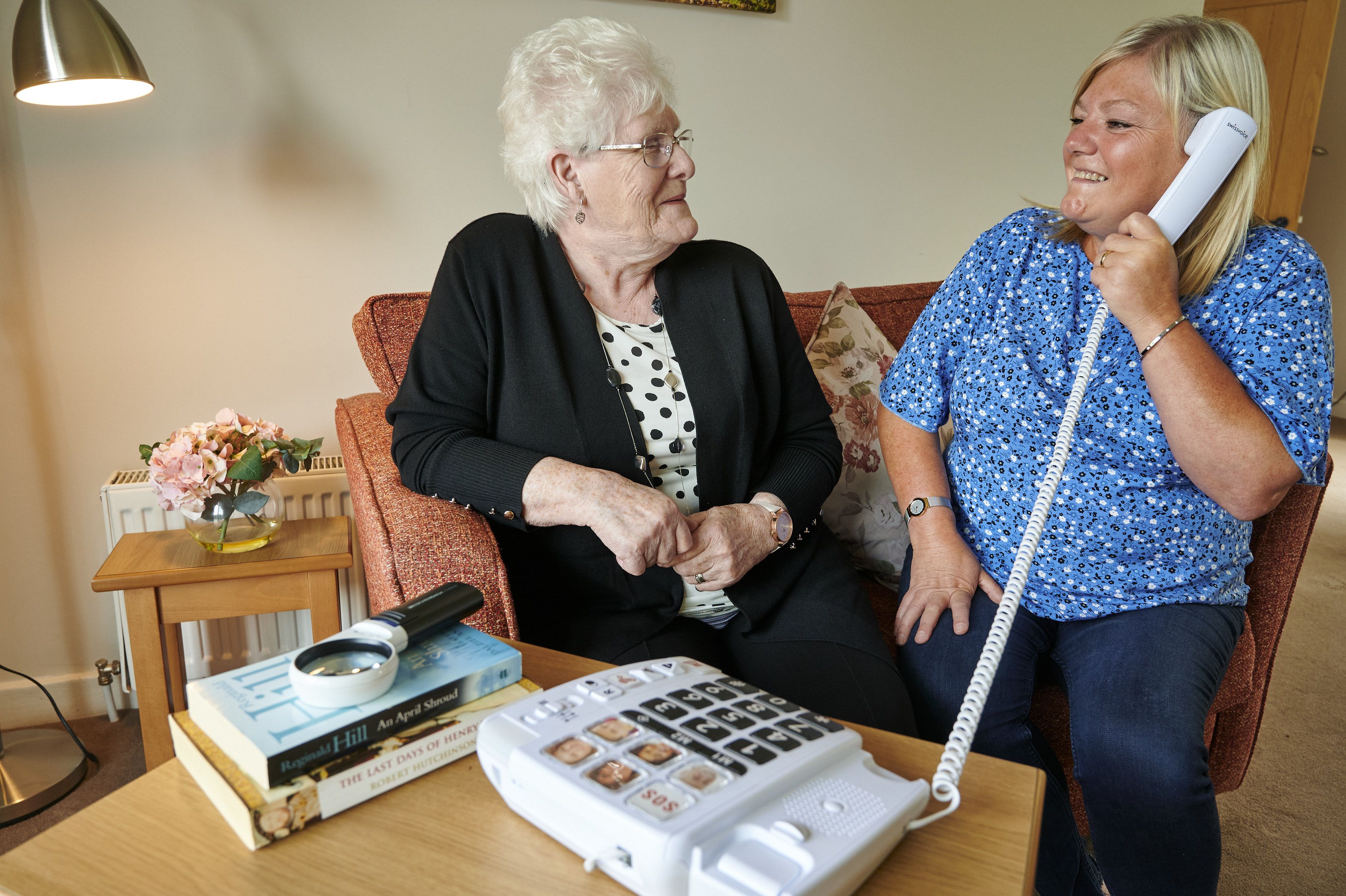 Veteran Anne Bell and Rehabilitation Officer Fiona McCormick on phone Two women sit smiling on the sofa. The younger lady on the right is making a telephone call.