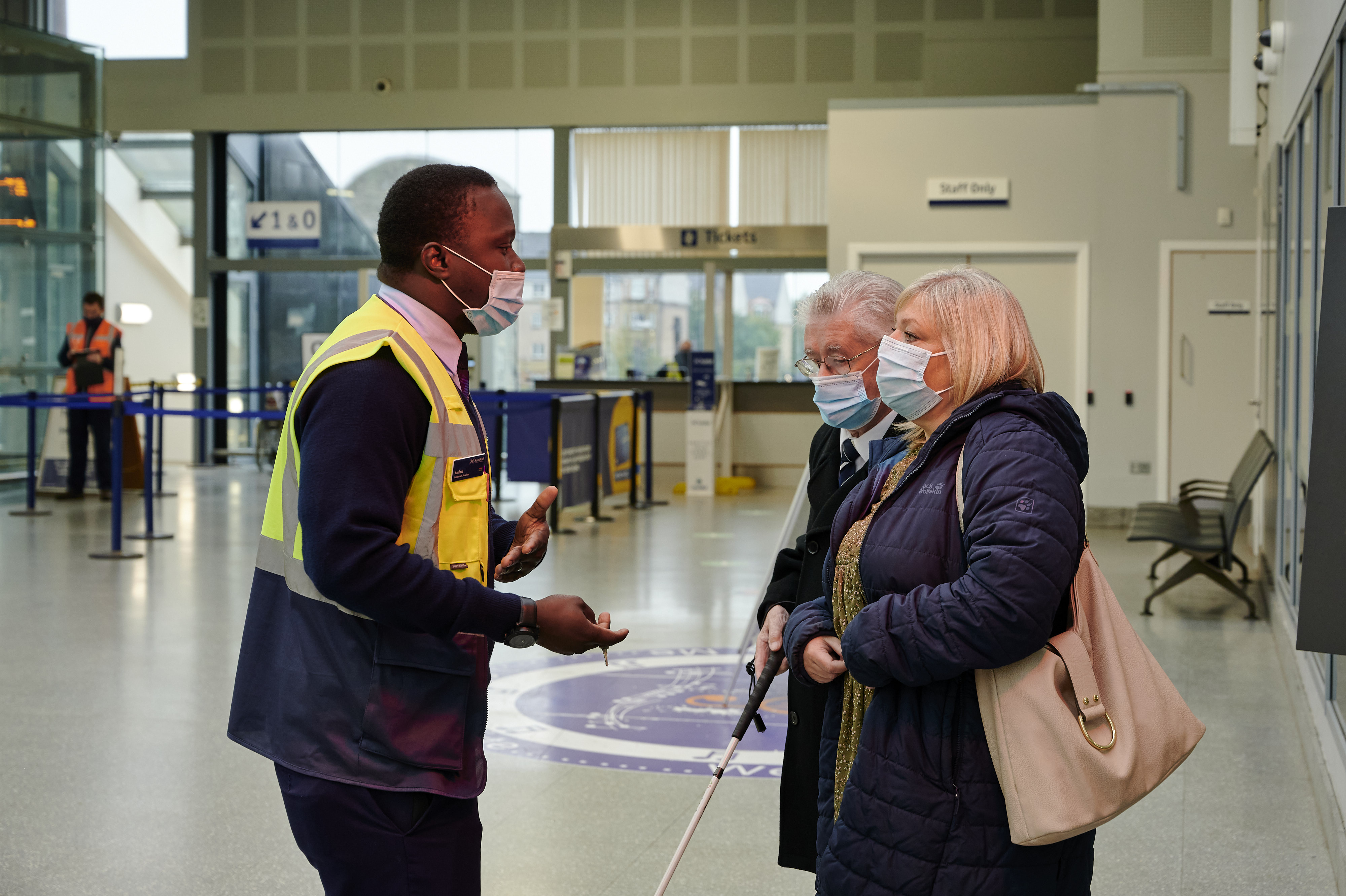 A rail worker assisting a visually impaired person with long cane and their companion in a train station in Scotland