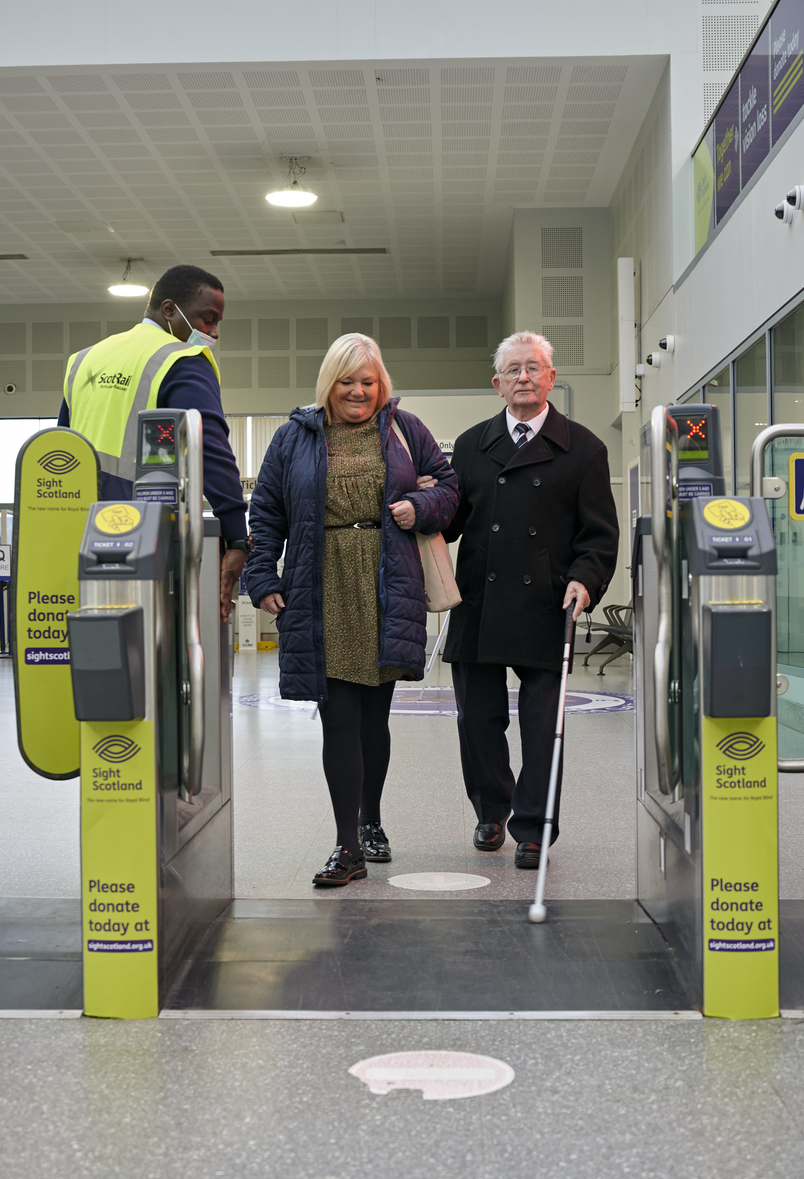 An individual with sight loss and their companion navigating the accessible gates assisted by a staff member in railway station