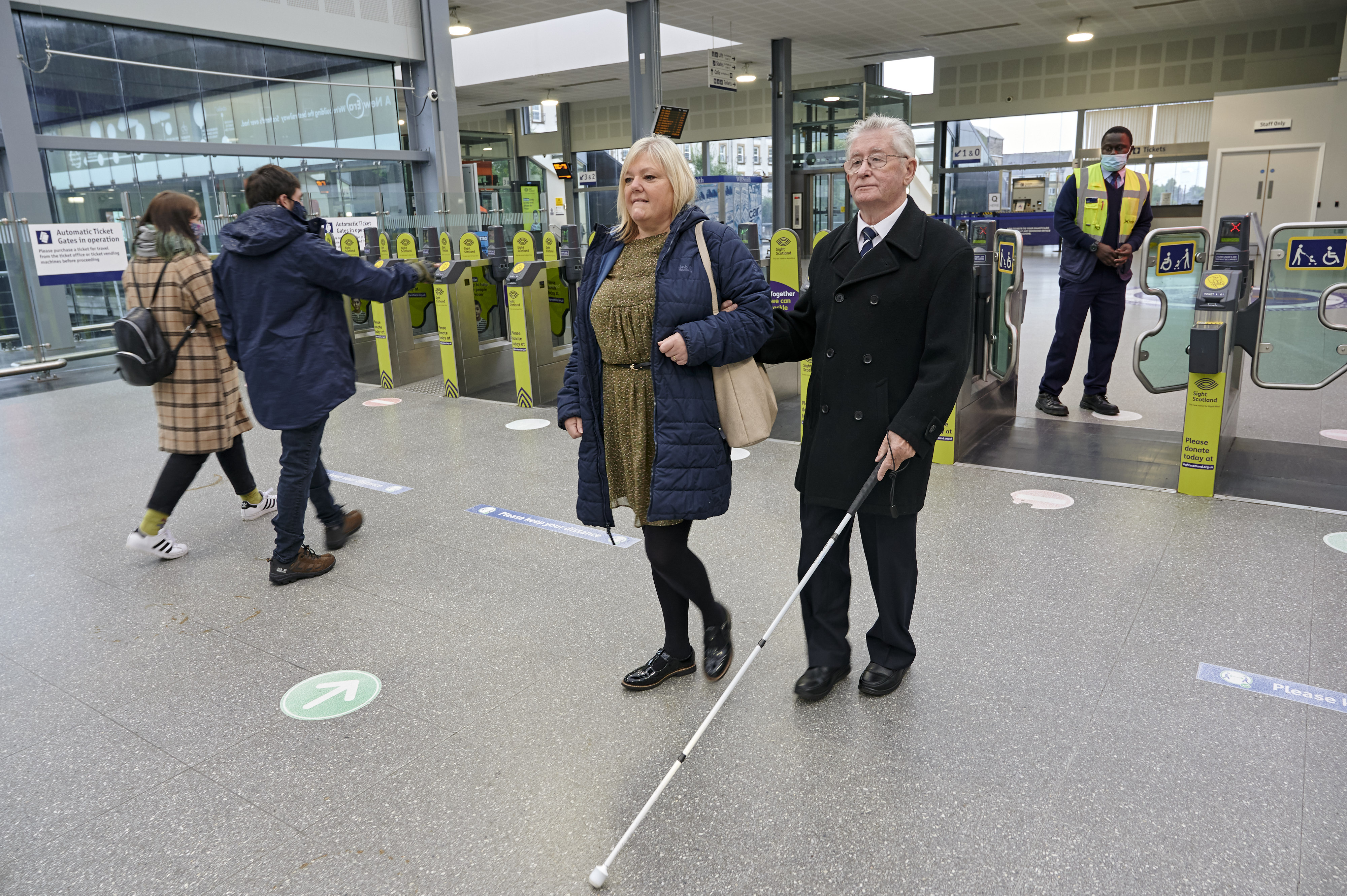 A person with sight loss and their companion walking with a long cane in railways station