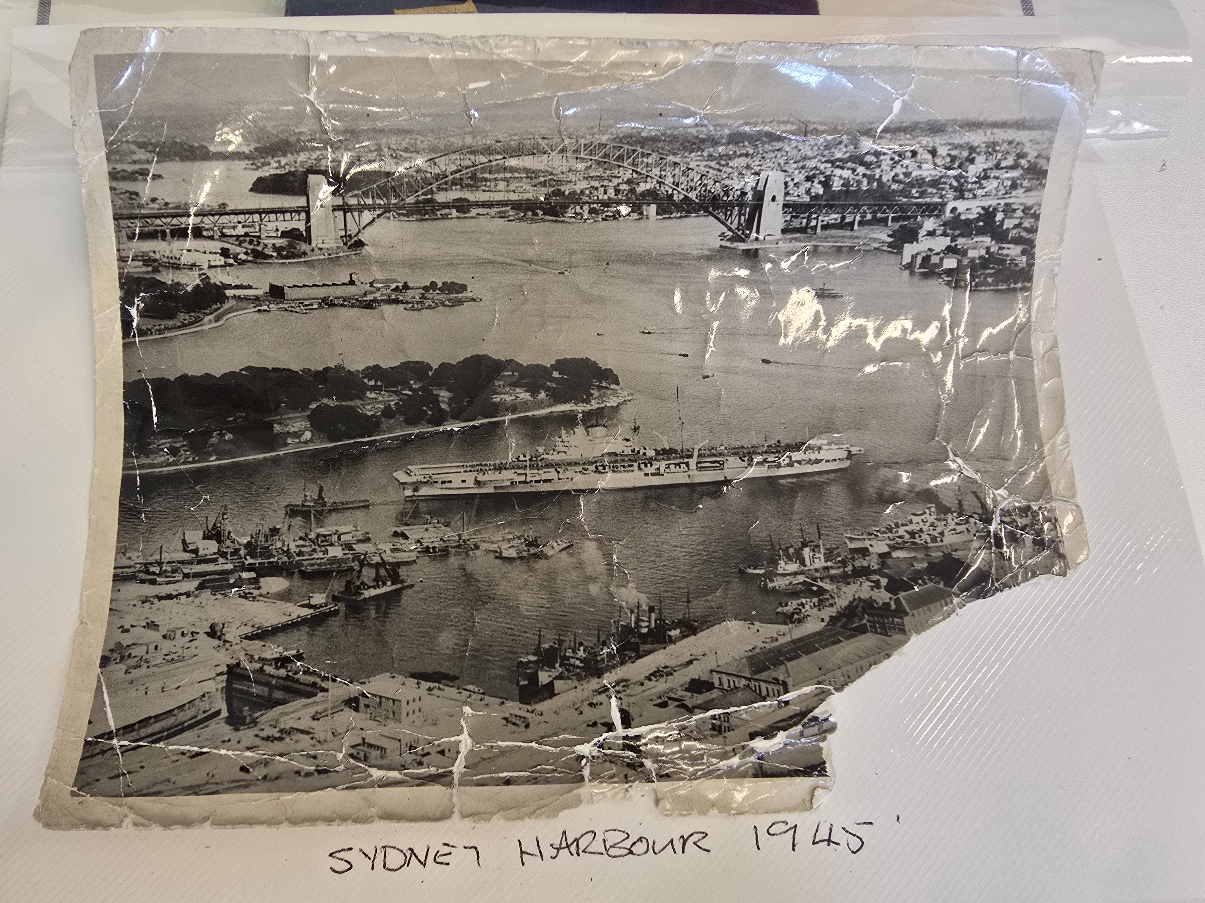 A black and white photo showing an aerial view of a harbour with a bridge in the background and a large boat docking. Hand-written below the photo is the text &quot;Sydney Harbour 1945&quot;