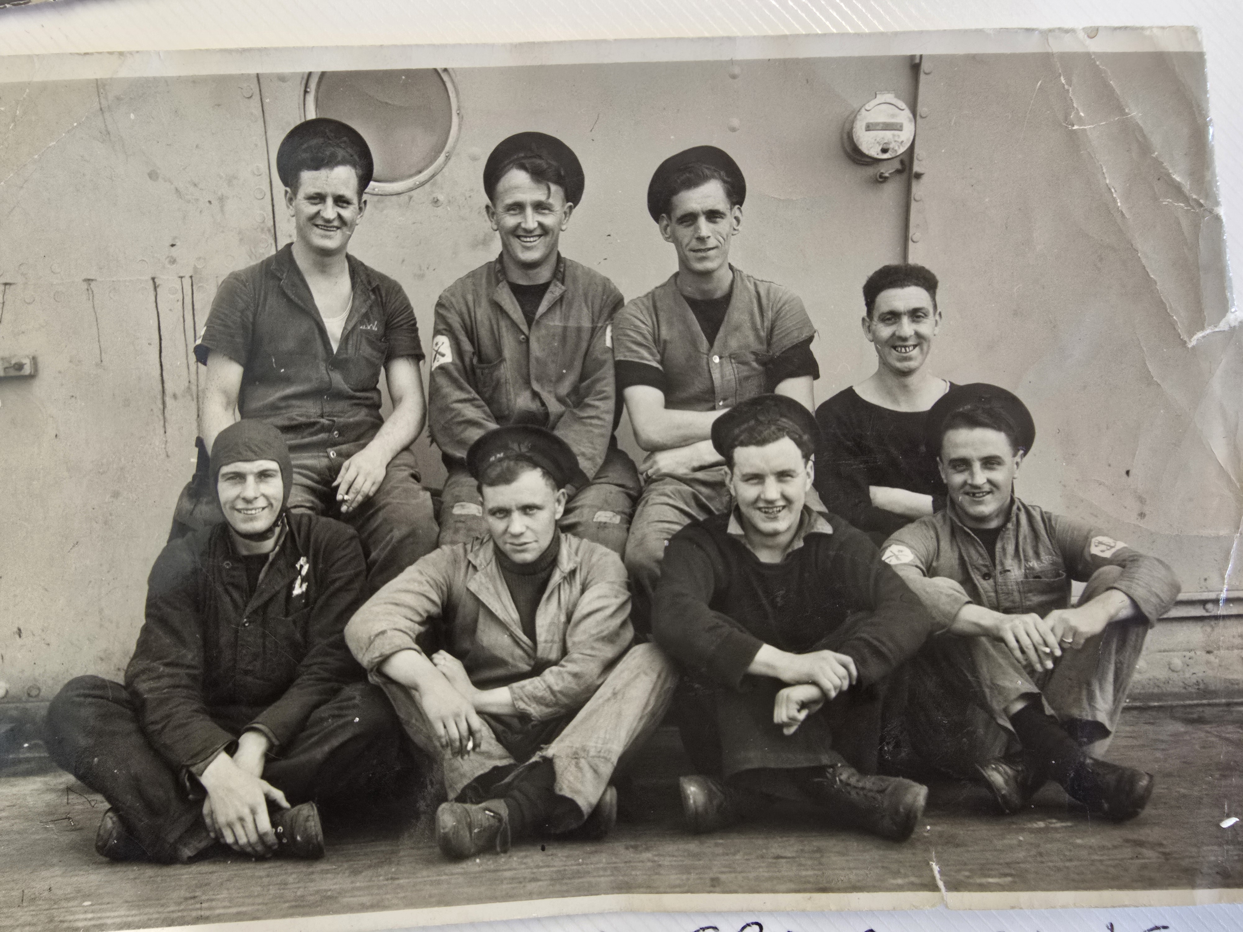 A black and white photo of a group of men sit on the floor facing the camera and smiling, they are wearing navy uniforms and some wear a beret.