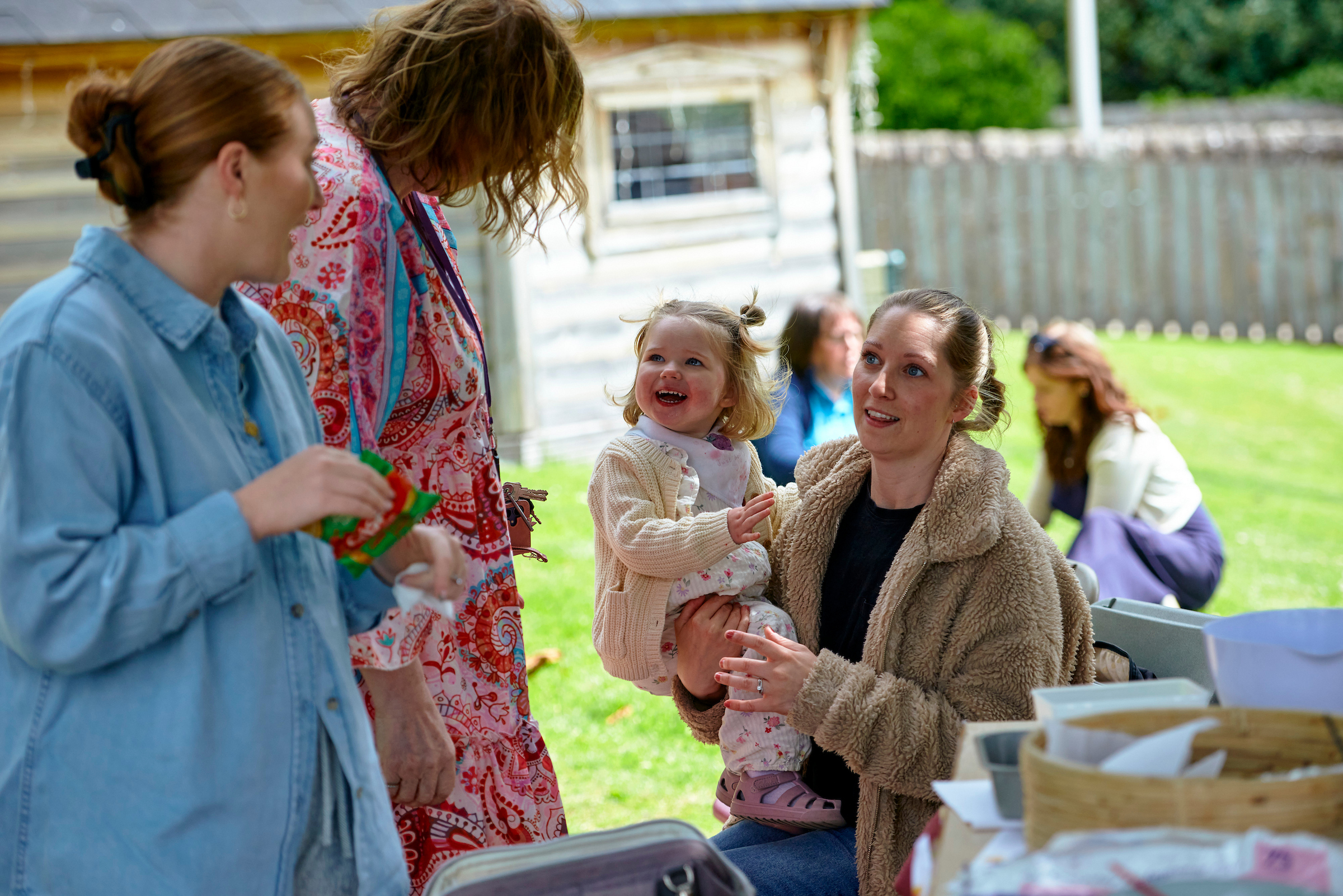 Two women stand outside talking to a woman who is seated and holding a very young smiling girl.
