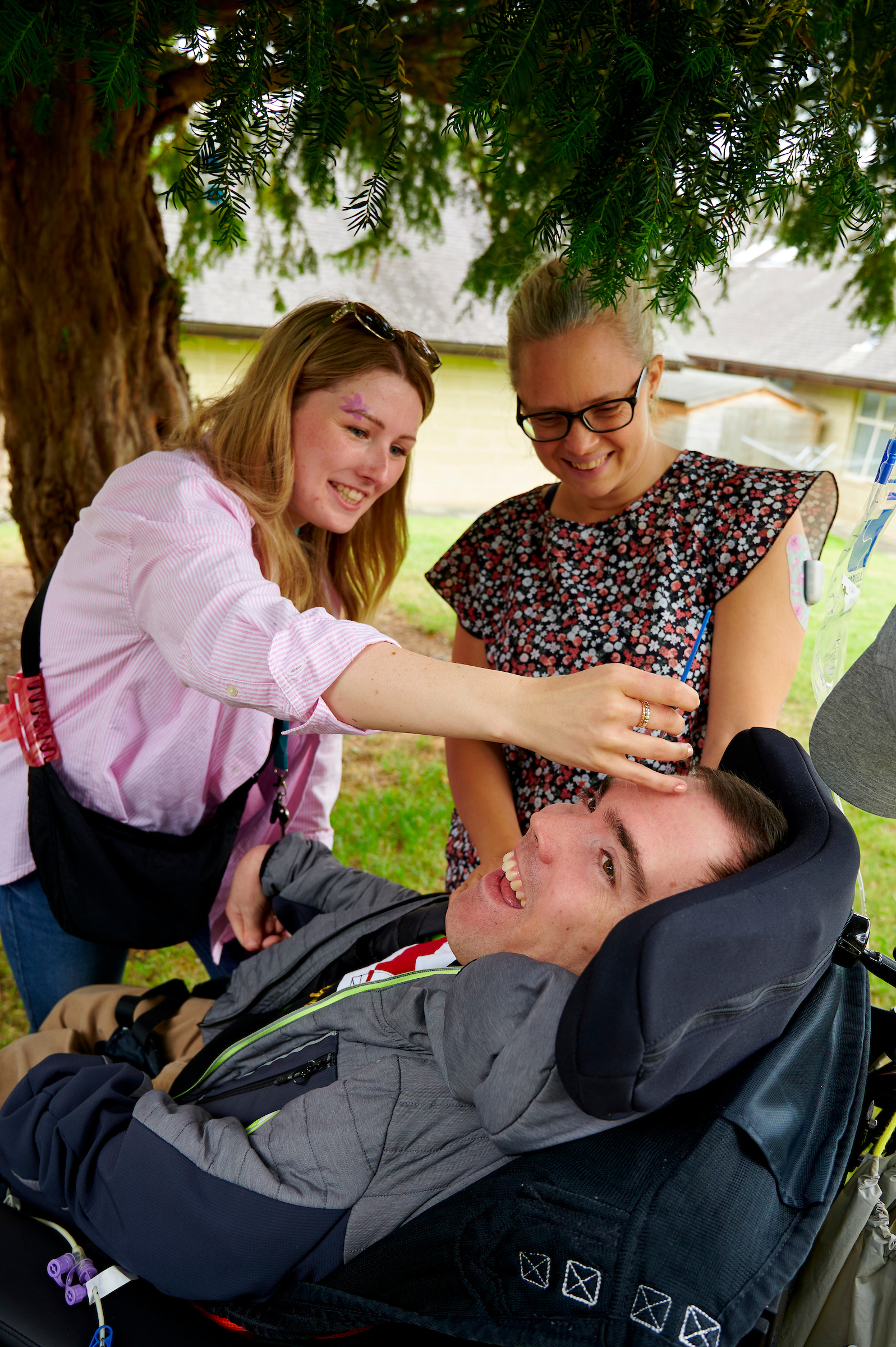 Two woman smile at a young boy in a wheelchair as he gets his face painted.