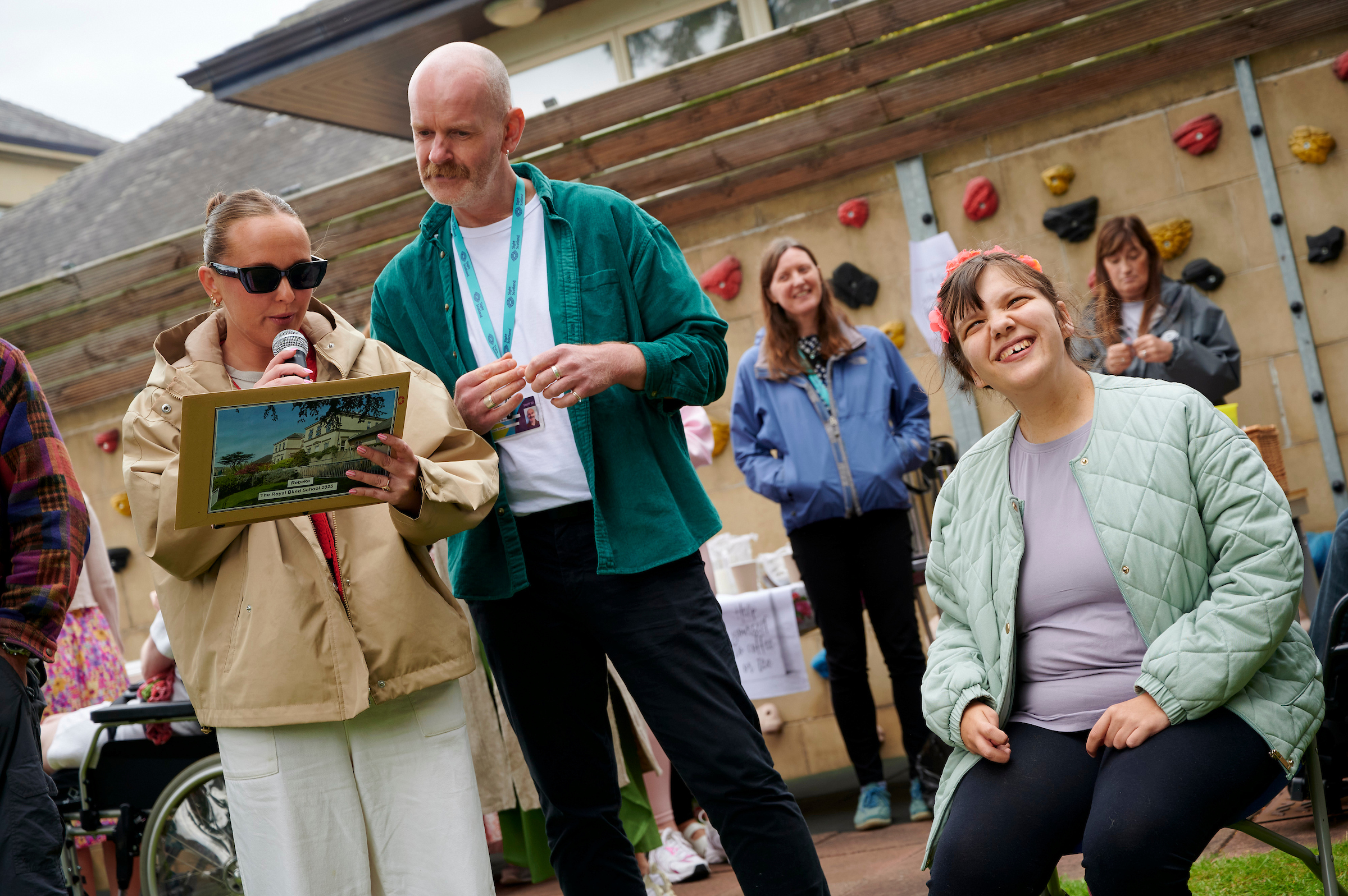 A woman in sunglasses and a man in a green jacket stand outside in front of a crowd, reading from a book, as a girl, Rebecca, sits beside them smiling.