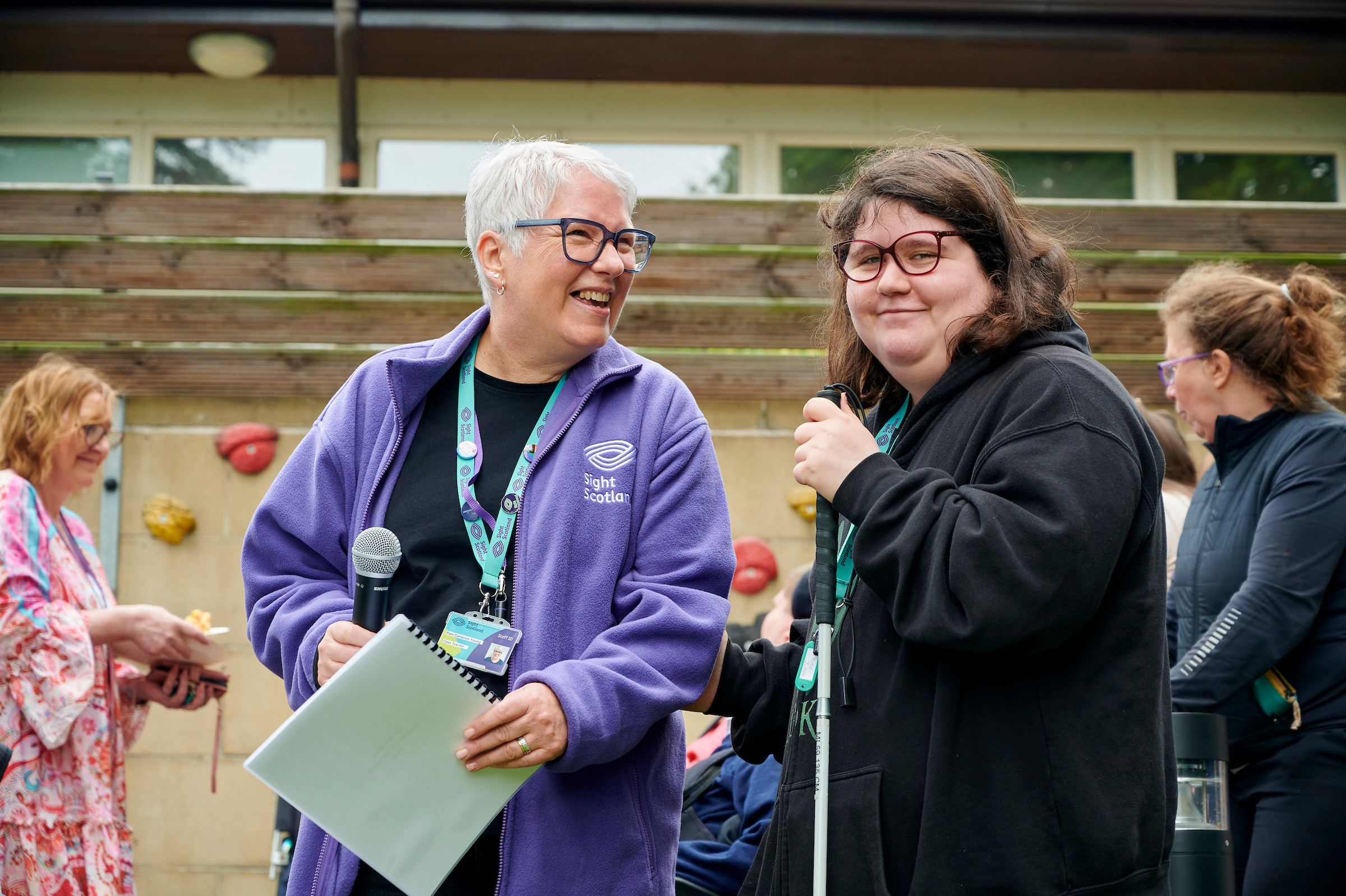 An older woman with short grey hair wears a purple Sight Scotland fleece and stands next to Jemima who wears red glasses and holds a cane