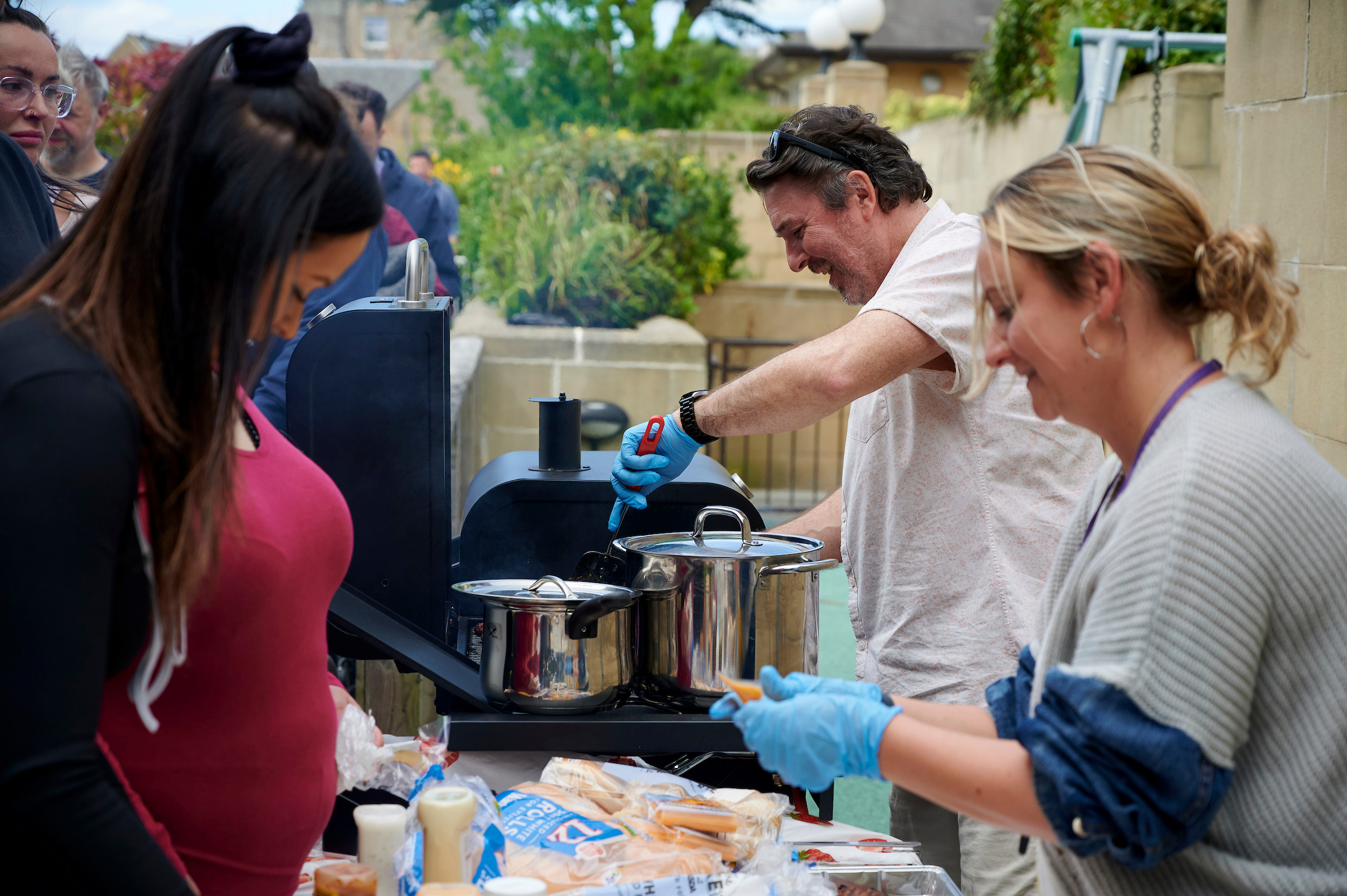 A man stands behind a barbeque grill and a woman next to him wears blue gloves as she serves people burger buns