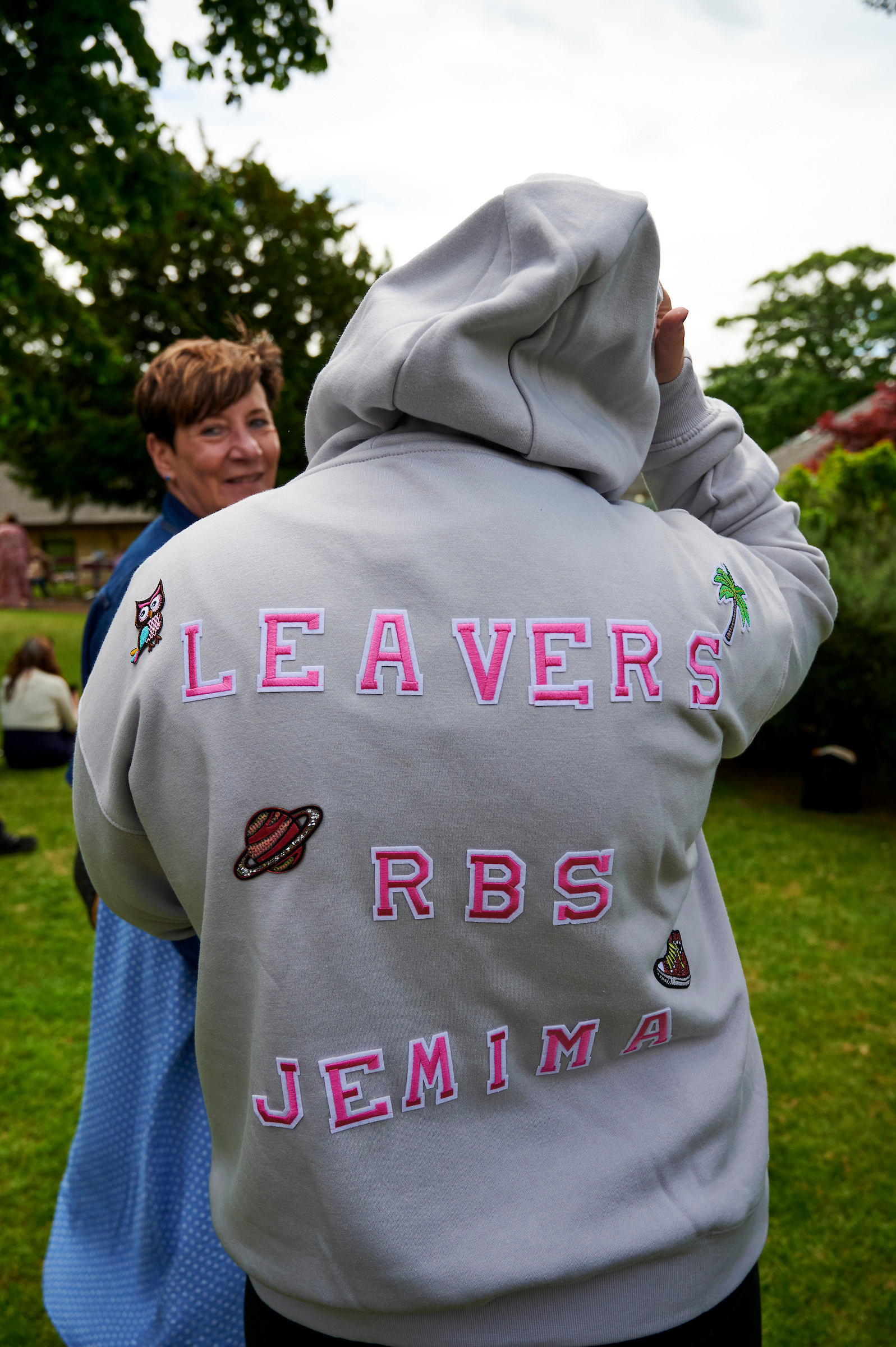 A person wears a light grey hooded jumper, the back emblazoned with the words 'LEAVERS RBS JEMIMA' in bright pink lettering