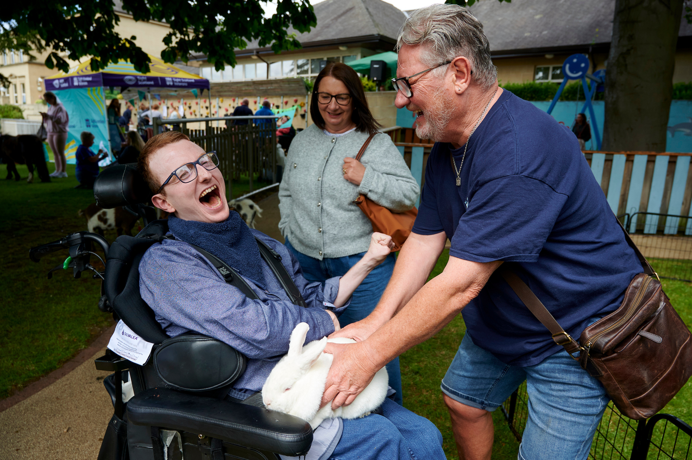 A young man in a wheelchair smiles widely as an older man holds a white rabbit in his lap