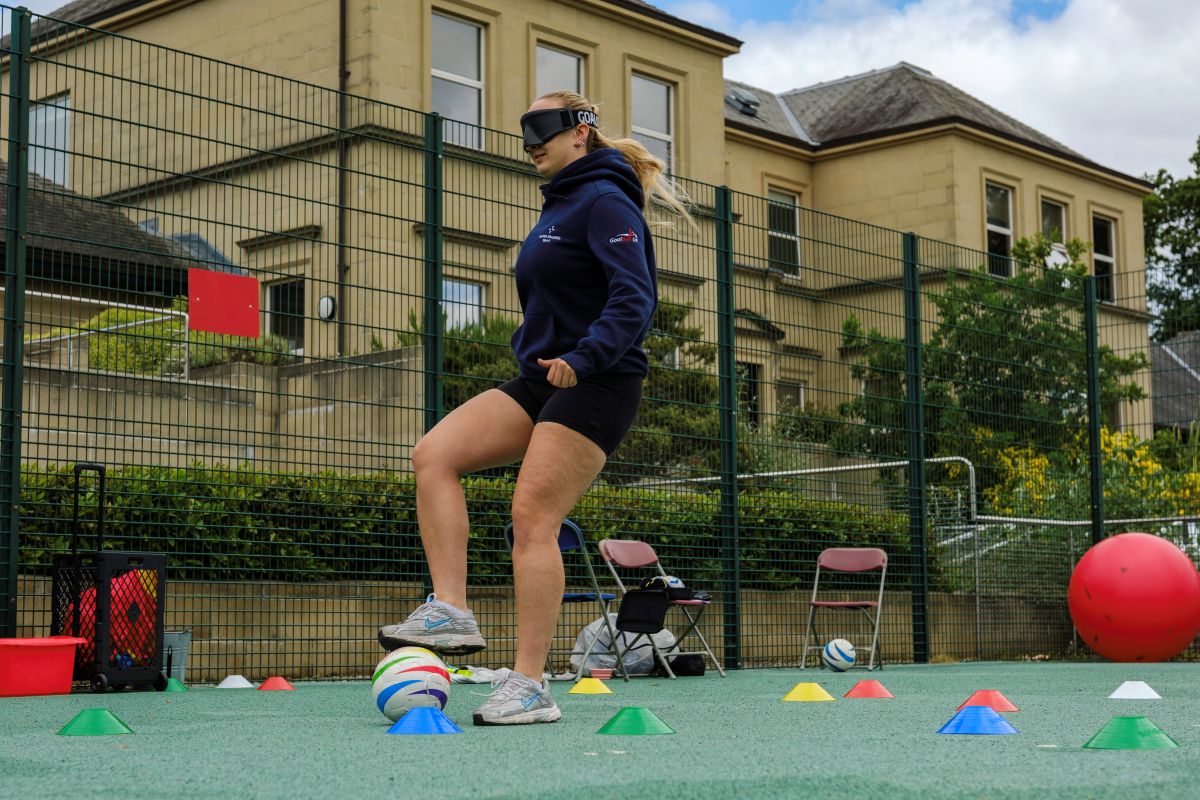 Sam Gough wears large dark sim specs and plays blind football on the pitch, kicking the ball between cones on the ground