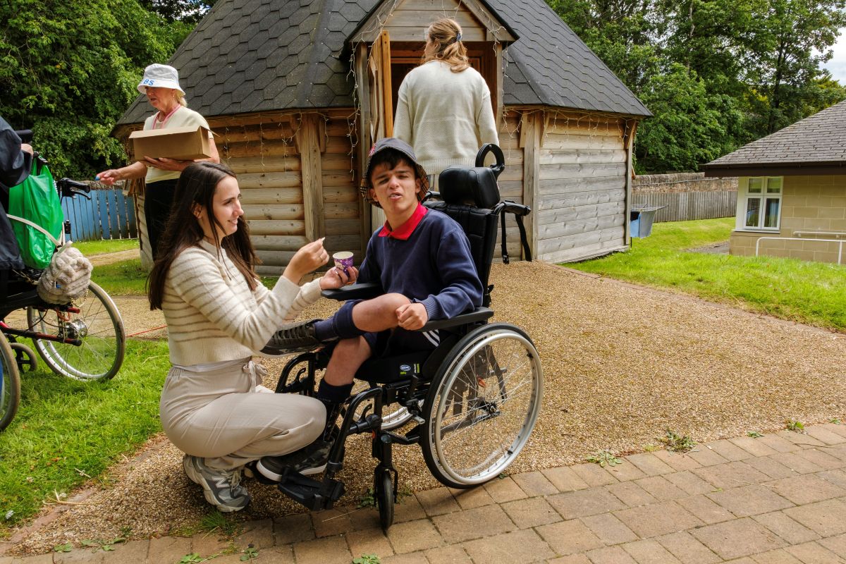 A pupil seated in a wheelchair outside in the school gardens makes a face as a woman crouched beside him offers him ice cream