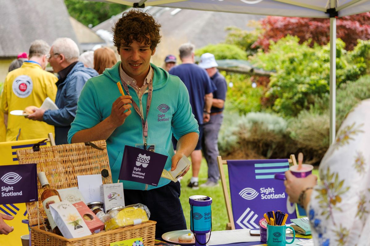 Supporter Fundraiser Sam stands behind a stand with Sight Scotland branded items displayed and a large hamper, he wears a green Sight Scotland jumper at the Royal Blind School Family Fun Day 2026
