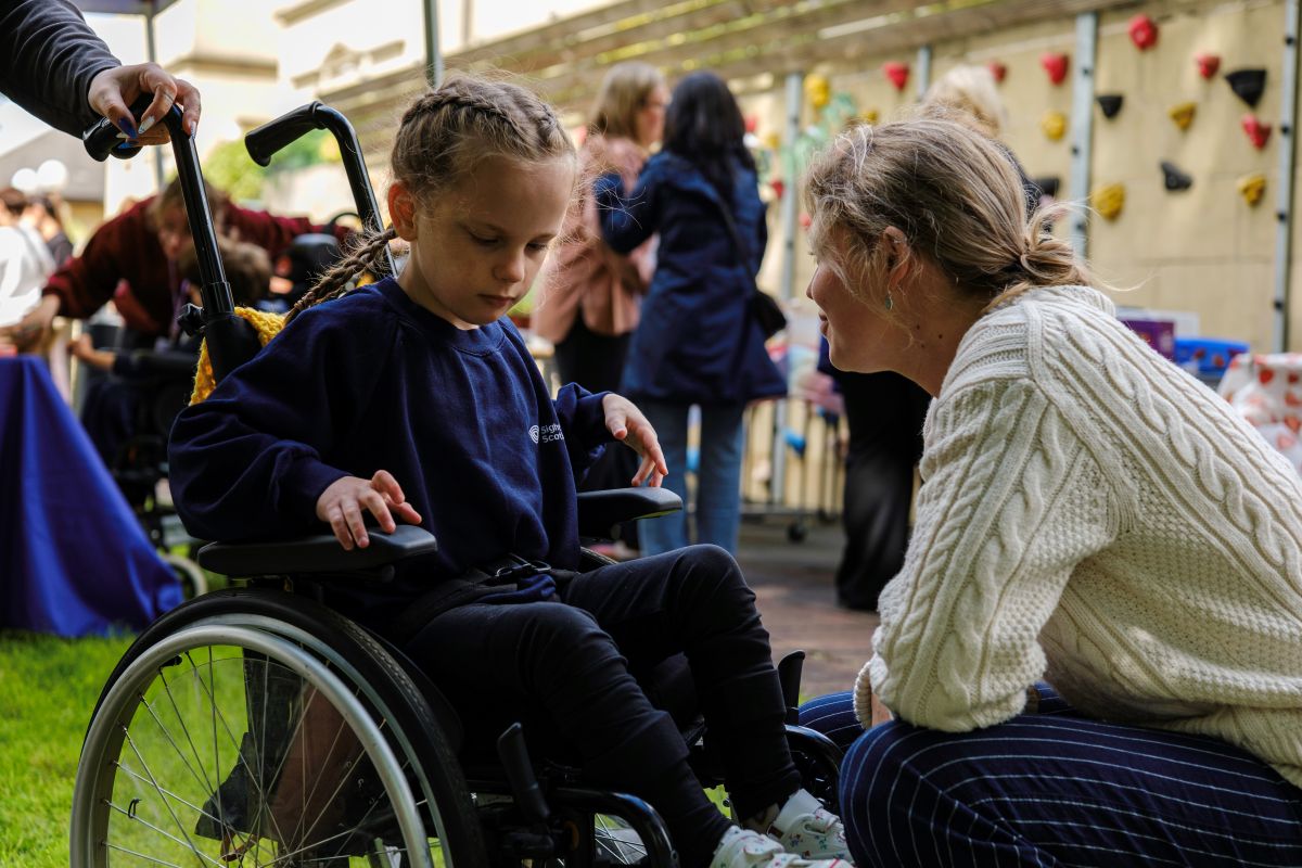 A woman in a white knitted jumper crouches down to talk to a small girl in a wheelchair at the Royal Blind School Family Fun Day 2026
