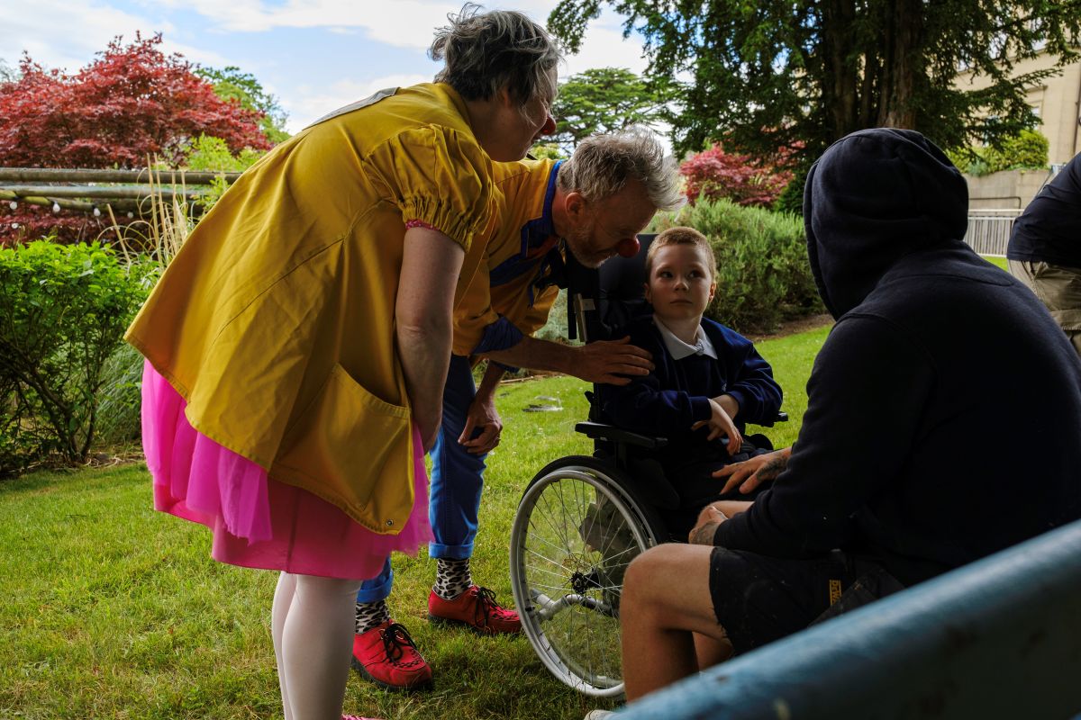 Two Clowndoctors, an older man and woman who are dressed in brightly coloured clothing and wearing novelty red noses, talk to a boy in a wheelchair at the Royal Blind School Family Fun Day 2026