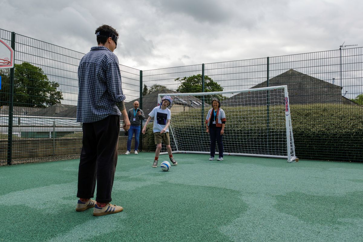 A group of people play blind football on a pitch, standing in front of a goal, one wears large dark glasses called Sim Specs, at the Royal Blind School Family Fun Day 2026