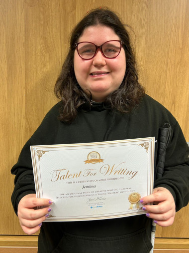 Jemima stands smiling in front of a wooden wall, holding a “Talent for Writing” certificate of merit. She wears red-framed glasses and a black hoodie. The certificate celebrates her original creative writing piece, selected for publication in a Young Writers’ anthology. Jemima has light purple painted nails and holds the certificate proudly with both hands.