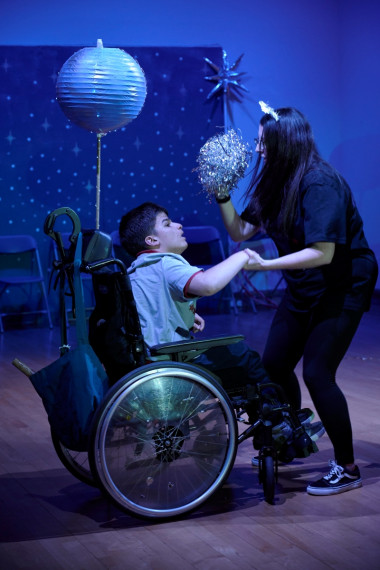 On a darkly lit stage, dances a woman in black waving a sparkly pom-pom and holding the hand of a young boy who sits in a wheelchair