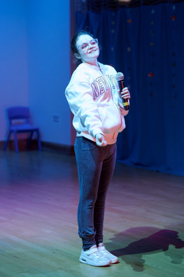 A young girl stands on-stage wearing a pink hoodie and jeans, she holds a microphone as she sings a brilliant solo to open the show
