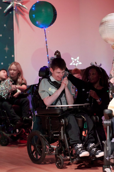 A boy on-stage in a wheelchair smiles widely and claps his hands, a sparkly balloon behind him, surrounded by other performers dressed for the show