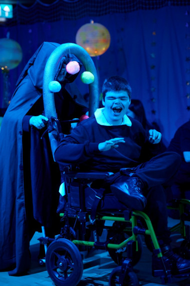 A boy in a space-themed decorated wheelchair grins widely whilst dancing to music, a person in a long black hooded robe behind him