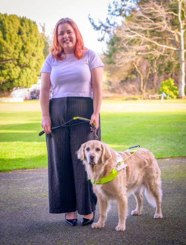 Lucy Edwards has long ginger hair and stands outside in a sunny garden with her guide dog, a golden retriever.
