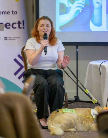 Lucy Edwards speaks to a crowd of people at the Connect event, she is sitting down and holding a microphone to speak into, her guide dog lying at her feet.