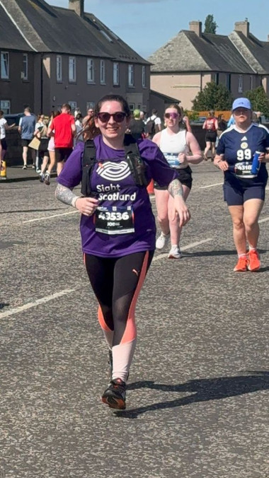 Jodie wears a purple Sight Scotland t-shirt and running gear as she runs down a busy street with other runners whilst completing the Half Marathon