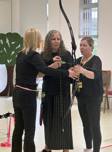 Sandi wearing black, holding an archery bow and arrow, standing in the middle of two women who are providing guidance