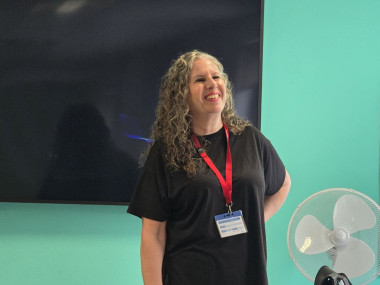 Sandi has curly grey hair, is wearing a black tshirt and red lanyard, smiling