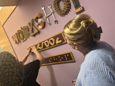 Sandi is touching a tactile signage board for a woodwork workshop above a braille sign