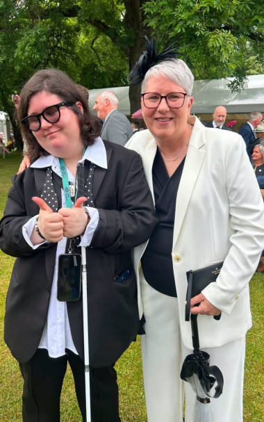 Jemima and Pam pose together at the Royal Garden Party. Jemima is wearing a black blazer over a white shirt with a black polka-dot tie and holding a white cane while giving two thumbs up. Pam is wearing a white trouser suit over a dark top, dark-framed glasses, and a black fascinator. Behind them are white marquee tents, and other guests in formal attire.
