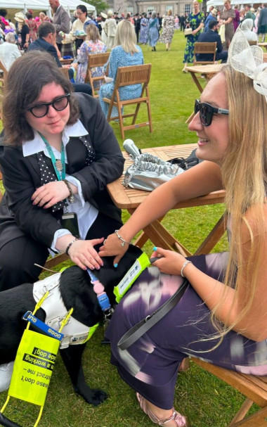 Jemima and Sam seated at a wooden table, reaching down to stroke Helga in her Guide Dogs harness.  Wooden chairs, marquees, and formally dressed guests fill the lawn behind them. Jemima is wearing a black blazer over a white shirt with a black polka-dot tie. Sam is wearing a dark purple patterned dress, sunglasses, and a light-coloured fascinator with loops and feather-like piece. 