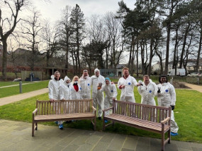 A group of people in white painting suits stand outside behind two freshly sanded wooden benches, thumbs up to the camera