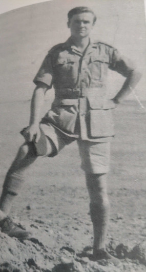 A black and white photo of a young man in military khaki jacket and long shorts, standing on sand, with his foot on a mount, his hand on his hip.