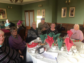 Five members sit at a table with Mick stood behind holding a cornet