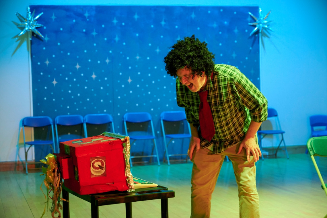 On stage, a man in a comic black wig screams at a sparkly decorated pretend computer, behind him a backdrop of stars.
