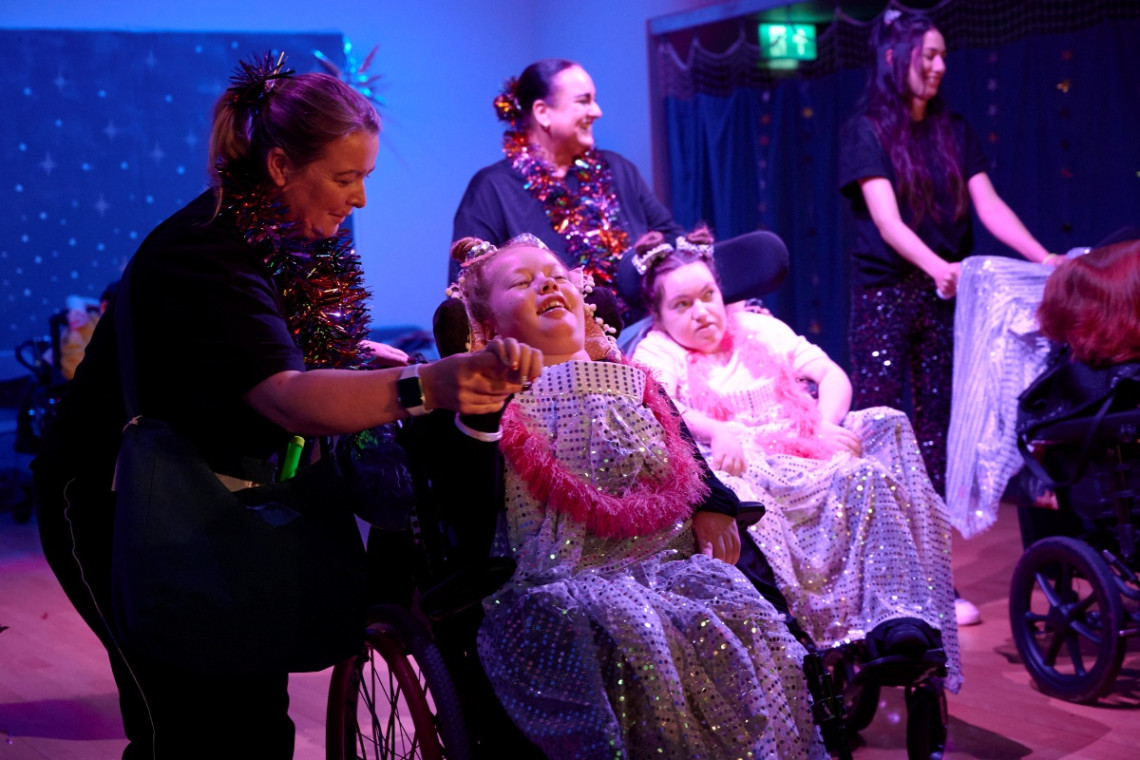 Two young girls in wheelchairs on a colourful stage, adorned with sparkly and starry costumes, alongside their teachers who wear tinsel around their necks