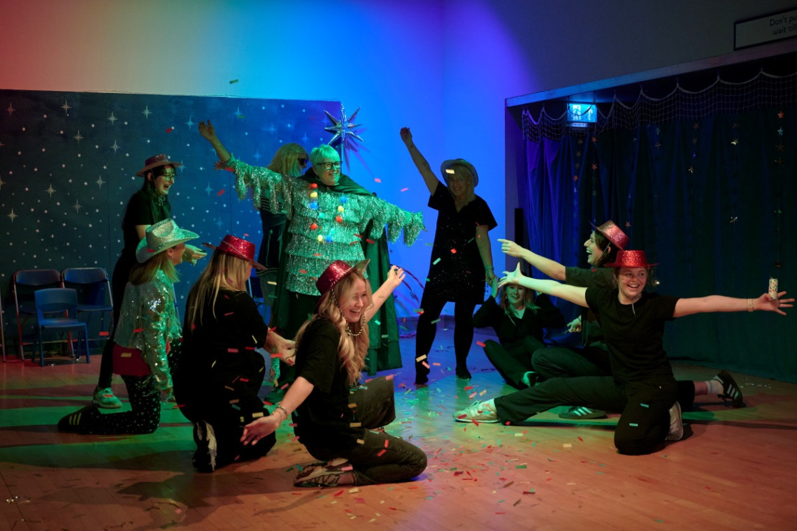 QTVI teacher, Pam, stands on-stage in the centre of a circle of people dressed in a sparkly silver Christmas tree outfit, the dancers surrounding her all wear red sparkly hats and are doing jazz hands