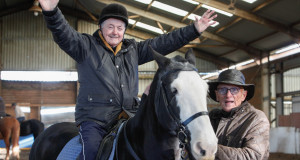 A veteran sits on a horse with arms outstretched while another veteran stands next to them