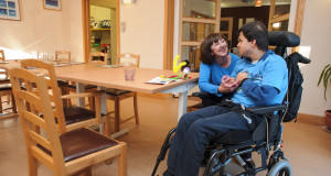 Pupil seated in wheelchair at dining room table smiles at care staff member