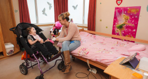 Pupil in wheelchair smiles at her mum who is seated on a bed with a pink duvet cover