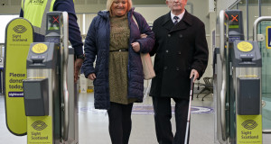 An individual with sight loss and their companion navigating the accessible gates assisted by a staff member in railway station