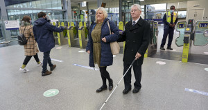 A person with sight loss and their companion walking with a long cane in railways station