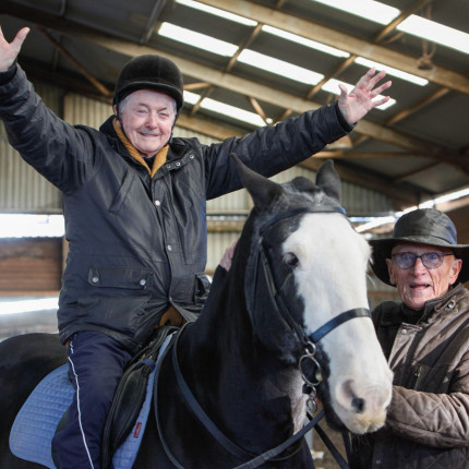 A veteran sits on a horse with arms outstretched while another veteran stands next to them
