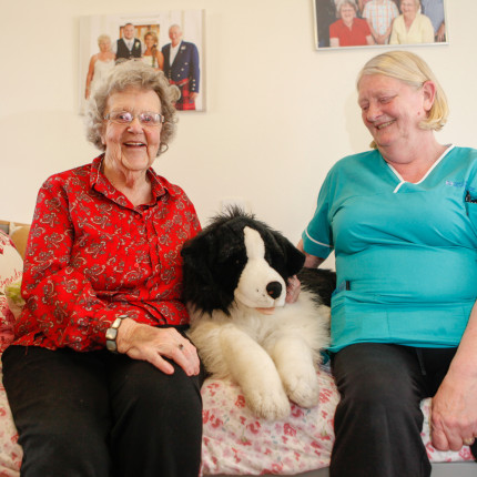 Braeside House resident and staff member sit on resident's bed with large stuffed dog toy between them